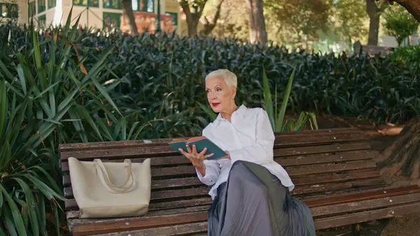 緑の公園で本を楽しんでいる引退したモデル 穏やかな朝だけで屋外で休む周りを見回して落ち着いた女性 庭で読書文学を破るスタイリッシュなシニアビジネスマン ロイヤリティフリーのストック画像