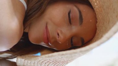 Hasır şapkalı esmer, gülümseyen yüz portresiyle güneşli bir kasabaya poz veriyor. Güneş ışığının tadını çıkaran muhteşem bir model dikey odaklı yakın çekim. Yaz tatilinde siesta yapan genç ve lüks bir kadın.