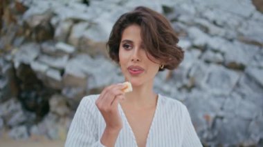 Deniz kıyısındaki kayalıklarda çörek yiyen güzel kız. Güzel bir kadın kameraya bakar ve dışarıda sağlıklı atıştırmalıkların tadını çıkarır. Plaj pikniğinde poz veren mutlu model. Gülümseyen dişi denizde rahatlar.