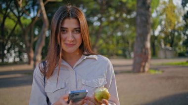 Yeşil güneşli parkta elinde elma tutan akıllı telefonlu nişanlı bir kadın. Güzel doğadaki mutlu kadın telefon numarasının portresi. Gelişigüzel yaşam tarzını yansıtan teknolojiyle yaz tatili.