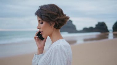 Harika esmer, konuşan cep telefonlu yakın plan. Kasvetli sahilde yürüyen güzel bir kız iş görüşmesi yapıyor. Kendine güvenen çekici kadın okyanus dalgalarında akıllı telefonla iletişim kuruyor. Seyahat yaşam tarzı konsepti
