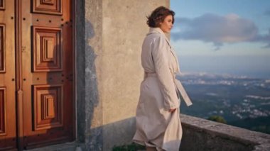 Rüzgarlı bir akşamda şehir manzarası seyreden muhteşem bir kadın. Düşünceli bayan, tarihi mimariye huzur içinde yansıyor. Trençkotla poz veren şık bayan gezgin güzel bulutlu şehir manzarasının tadını çıkarıyor.