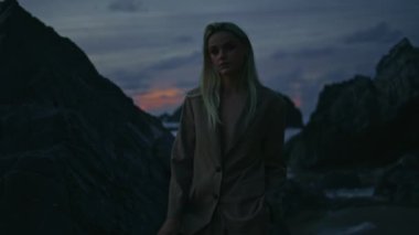 Karanlık bir akşamda manken kız sahili. Parlayan ışıkta taş pozu veren seksi kendine güvenen sarışın. Bulutlu deniz kıyısında bir kadın silueti. Okyanus kumsalı taşlarına ateş eden zarif sarışın görünümlü kamera.