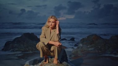 Akşamki okyanus dalgalarında kayalıklarda oturan çekici bir model. Gün batımında açık renkli poz veren, sarı saçlı bir kadın. Şık takım elbiseli seksi, şık bir kız. Cazibeli çekim seansında kameraya bak.