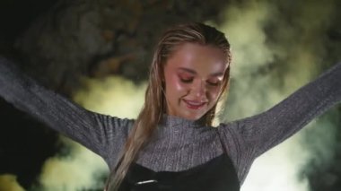 Duman bombası bulutu içinde dans eden mutlu kadın. Çekici kız projektör ışığında müzik dinlemekten zevk alıyor. Neşeli ve çekici bir bayan, karanlık arka planda festivale ayak uyduruyor.