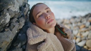 Sakin bir kadın okyanusta yalnızlığın tadını çıkarıyor. Kış plajında dinlenen huzurlu kız şık bir ceketle kameraya bakıyor. Yumuşak deniz taşlarına poz veren rahat bir model. Hafta sonu gezintisi. 