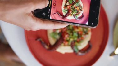 Kadın eli restoran masasında cep telefonuyla lezzetli bir yemeğin fotoğrafını çekiyor. Kimliği belirsiz kişi dijital akıllı telefondan dikey görüntü alıyor. Müşteri kafede yemeğin tadını çıkarıyor.. 