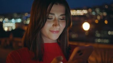 Güzel ışıklandırılmış şehir manzarasında akıllı telefon kullanan akşam kızı. Gülümseyen kadın gece yürüyüşünde sosyal medyada mesajları kontrol ediyor. Güzel bir bayan yalnız başına internette geziniyor.