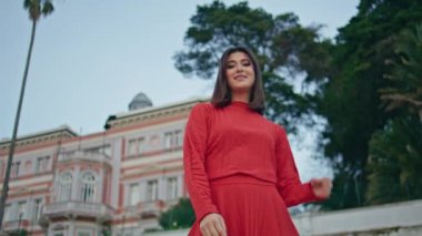 Güzel bayan şehir manzarasının portresi. Kırmızı elbiseli, gülümseyen sakin kadın kameraya bakıyor ve bahar gökyüzü doğasında esmer saçlara dokunuyor. Kasabanın bahçesine hayran olan mutlu manken yalnız zaman geçirmeyi seviyor. Şık kız. 