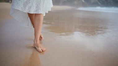 Okyanus kıyısında ayak izleri bırakan kadın bacakları. Kaygısız kız ıslak kumda yürüyor ve huzur dolu sahil doğasının tadını çıkarıyor. Tanınmayan romantik bayan tropikal sahillerde geziniyor. İnce ayaklar kayalık adaya gidiyor