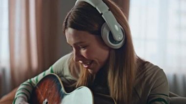 Mutlu kadın dairede kulaklıkla gitar çalma pratiği yapıyor. Gülümseyen kadın gitarist evdeki kanepede akustik enstrüman ile melodi besteliyor. Müzik eğitimi veren yaratıcı kız müzisyen.