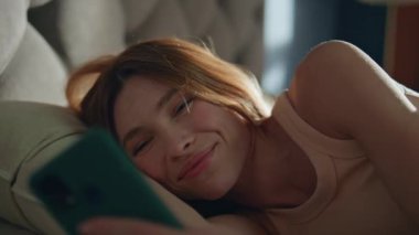 Cep telefonu mesajı okuyan uykulu kadın güneşli bir sabah uyanıyor. Rahat gülümseyen kız rahat bir yatakta uzanmış akıllı telefon ekranına bakıyor. Güzel tembel bayan rahat yatak odasında tek başına uyuyor..