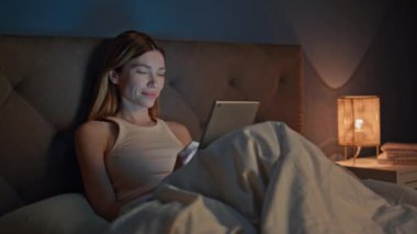 Gecenin geç saatlerinde rahat bir yatakta tablet giyen bir kadın. Gülümseyen çekici kız karanlık yatak odasındaki bilgisayardan sosyal medyayı izliyor. Yatma vakti dijital aletlerle uğraşan rahat bir bayan. Bağımlılık kavramı.