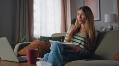 Öğrencilerin evdeki kanepede deftere yazı yazma dersleri var. Modern apartmandaki bilgisayarlı video konferansında internet öğretmenine gülümseyen akıllı kadın. Kendine güvenen kız müşterinin yaptığı işleri not ediyor..