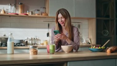 Ev hanımı hafta sonu sabahı mutfakta cep telefonuyla mesajlaşıyor. Gülümseyen kadın akıllı telefon ekranında mutfak tezgahında dikiliyor. Mutlu kız eve yemek pişirdikten sonra cep telefonunda mesaj okuyor.