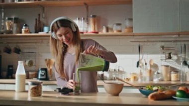 Dans eden kadın smoothie hazırlıyor rahat mutfakta kablosuz kulaklıkla müzik dinliyor. Kulaklıklı gülümseyen kız yeşil sebzeleri cama döküyor. Mutlu bayan dairede kahvaltı hazırlıyor..
