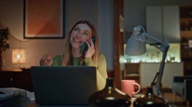 Bilgisayarlı cep telefonuyla konuşan pozitif bir uzaktan kumandalı. Yakın çekim. Gülümseyen çekici kadın cep telefonu konuşmasından hoşlanıyor. Karanlık dairede çalışıyor. Uzak iletişim
