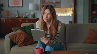 Kadın evdeki kanepede tablet ekranına dokunuyor. Çevrimiçi iş için bilgisayarını kullanan çekici sakin bir kız. Oturma odasında kahve fincanıyla oturuyor. Rahat internet müşterisi uzak alımları seçer.