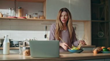 Modern mutfakta yemek pişiren kadın dizüstü bilgisayar ekranı arıyor. Ciddi bir iş kadını bilgisayardan sabah kahvaltısı hazırlıyor. Çekici ev hanımı salatayı karıştırıyor. Mutfakta granola yapıyor.
