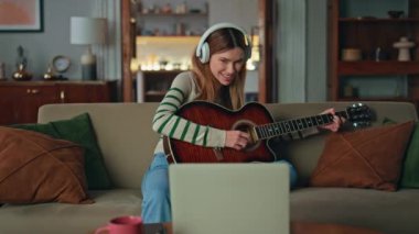 Oturma odasındaki kanepede gitar çalan gülümseyen kadın. Kulaklıklı mutlu bir bayan gitarist dizüstü bilgisayara bakan bir web kamerası kullanıyor. Olumlu müzisyen kız evde enstrümantal müzik dinlemeyi sever.