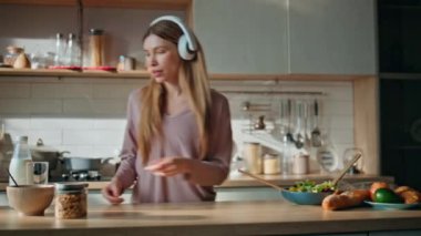 Kulaklıklı kadın açık mutfakta kahvaltı hazırlıyor. Kaygısız kız masada sebze salatası karıştırarak müzik dinliyor. Sağlıklı yemek hazırlayan mutlu bir kadın..