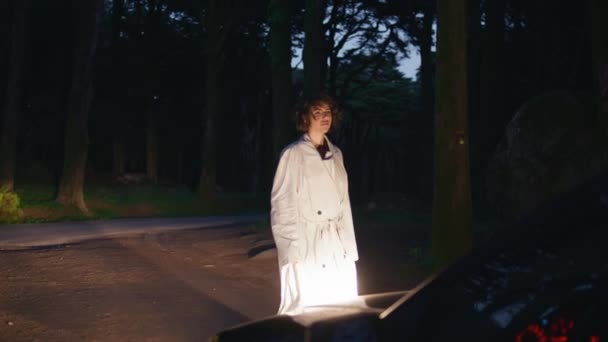 夜の森林道路の車のヘッドライトに照らされた女性 女性のシルエットは 暗い木に対してキャスト リラックスした女性が暗い自然の中で自動車の光に立って旅を熟考 ハッピー ブルネット — ストック動画