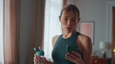 Spor salonundaki kız, sabah antrenmanından sonra akıllı telefon gibi görünüyor. Egzersiz yaptıktan sonra elinde su şişesi tutan ciddi yorgun sporcu kadın. Atletik kadın cep telefonunda antreman uygulaması.