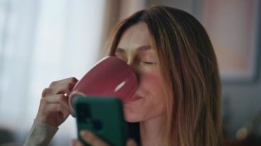 Portredeki kadın evde akıllı telefon haberleri izlerken kahve içiyor. Güzel, rahat bir kadın, yakın çekimde sıcak çay görünümlü cep telefonu ekranının keyfini çıkarıyor. Sakin kız hafta sonu cep telefonu uygulamasına göz atıyor
