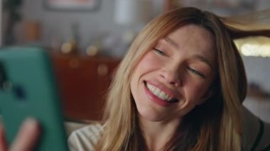 Oturma odasında cep telefonu tutan kız arkadaş videosu. Kapatın. Duygusal açıdan mutlu bir kadın akıllı telefonlu kamerayla konuşuyor ve evi dinlendiriyor. Gülümseyen güzel bayan çevrimiçi iletişimden hoşlanıyor.