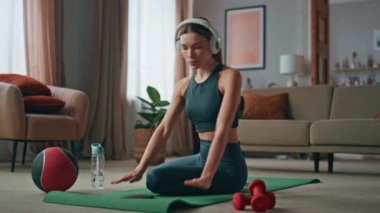 Spor kadını evdeki spor minderinde kablosuz kulaklık takıyor. Ciddi bir kadın günlük antrenmanlarda pilates antrenmanı yapıyor. Fit kız kulaklıkla müzik dinleyip kıvırıyor..