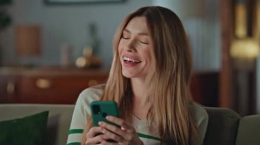 Evdeki oturma odasında cep telefonuyla mesajlaşan mutlu bir kız. Neşeli, genç bir kadın cep telefonu okuyor. Hoş bir mesaj, otururken gülümsüyor. Sevimli, çekici bir bayan erkek arkadaşına mesajla cevap veriyor..