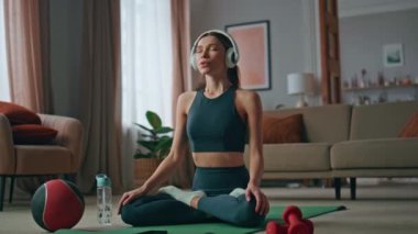 Evde kulaklık takan, Lotus pozisyonunda oturan ilham verici bir kadın. Sakin, kapalı gözlü kız oturma odasında spor minderinde yoga yapıyor. Kablosuz kulaklıkla ses meditasyonunu dinleyen sakin kadın yogi