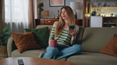 Kahve fincanıyla rahatlayan ve elinde akıllı telefon tutan bir kız. Evcil genç bayan sıcak içeceğin keyfini çıkarıyor. Evde rahat bir koltukta cep telefonuyla oturuyor. Çekici ev hanımı hafta sonu evde tek başına dinleniyor..