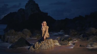 Gece sahilinde kayalıklarda oturan bir kadın. Karanlık okyanus kıyısında poz veren, takım elbiseli bir model. Alacakaranlıkta dergi için ateş eden güzel bir kadın. Yalnız sarışın kız yaz akşamı kendine sarılıyor. 