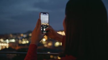 Kız turist akıllı telefondan şehir manzarasını aydınlatan bir fotoğraf çekiyor. Esmer kadın akşam panoramik kare arka görüşünde modern cep telefonu kullanarak fotoğraf çekiyor. Dijital cihazı tutan mutlu bayan