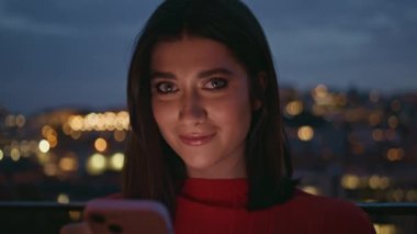 Gece portresi gülümseyen kadın terasta akıllı telefon tutuyor. Mutlu bayan aydınlatılmış şehir manzarasında sosyal medyayı kontrol ediyor. Neşeli romantik kız cep telefonu kullanarak online ileti oluşturuyor 