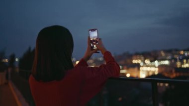 Bilinmeyen bir kadın, gece yakın plan akıllı telefon kullanarak şehir manzarasını fotoğraflıyor. Esmer kız aydınlatılmış kasaba manzarası çekiyor. Rahatlamış bir kadın karanlıkta meydanda tek başına fotoğraf çekiyor.