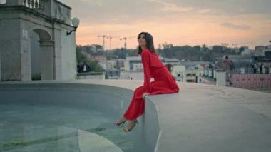 Dişi esmer dinlenme çeşmesi Sunset City 'de pansiyonerlik yapıyor. Kaygısız kadın suya bakıyor ve sakin bir akşamın tadını çıkarıyor. Mutlu romantik model kırmızı elbiseye dokunuyor. Güzel yaz parkında dinleniyor.