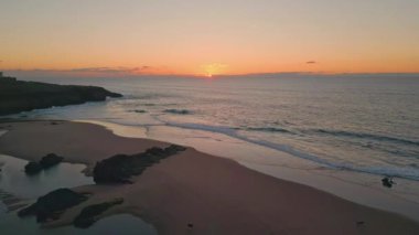 Gün batımı kayalık sahili aydınlatıyor. Sakin bir hava görüntüsüyle. Şafak, manzaralı sahile huzurlu bir renk kattı. Yumuşak dalgalanan dalgalar sakin sabah manzarasında kıyıları yıkıyor. Güzel gün doğumu gökyüzü manzarası.