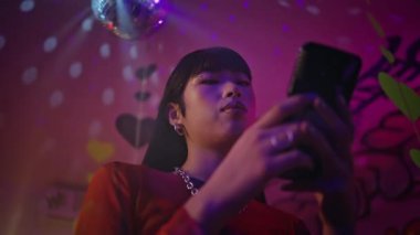 Neon Light Kulüp 'teki ciddi görünümlü akıllı telefon en alt görüntüde. Parlak makyajlı güzel Çinli kız gece disko partisinde telefonla mesajlaşıyor. Çağdaş kadın gece kulübünde mesajlaşıyor..
