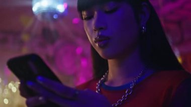 Şehir kulübünün ışıklarında akıllı telefona mesaj yazan gülümseyen bir kız. Neon disko partisinde sosyal medyada gezinen güzel Asyalı kadın. Genç esmer, gece hayatı etkinliğinde cep telefonu mesajı okuyor..