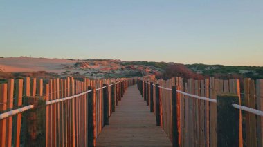 Gün batımında ahşap yol canlı kum tepelerinin arasından geçiyor. Akşam, doğa koruma sahasına sakin bir turuncu ışıltı saçıyor. Manzaralı yol iskelesi sahil şeridinde sakin bir açık hava kaçışı sunuyor..