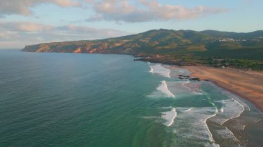Turkuaz dalgalar akşam güneşliğinde kumsalda dönüyor. Gün batımı kıyıları ve dağları kucaklıyor. Alacakaranlıkta el değmemiş okyanus kıyısının sakin manzarası. Engebeli arazinin deniz manzarası. Seyahat tatili.