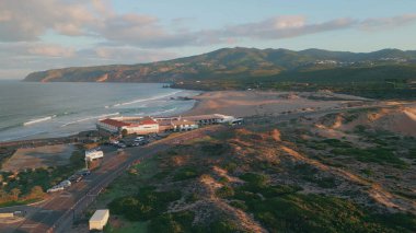 Gün batımında sakin sahil yolu. Karanlık çöktüğünde engebeli uçurumlara karşı kıyı kasabasının hava manzaralı yuvası. Altın saat, sakin okyanus ve sıcak kum tepeleri banyosu. Görkemli deniz dronu panoraması