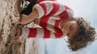 Okyanus kıyısında kum figürleri yapan sevimli çocuk. Odaklanmış kıvırcık çocuk dışarıda istirahat ediyor ve sonbahar günışığında dikey olarak plaj şatosu yapıyor. Kaygısız anaokulu çocuğu bilinmeyen bir anneyle oynuyor..