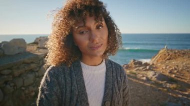 Deniz uçurumunda poz veren yumuşak esmer doğa portresinin keyfini çıkarıyor. Muhteşem doğal makyaj modeli fırtınalı denizin önündeki kameraya benziyor.