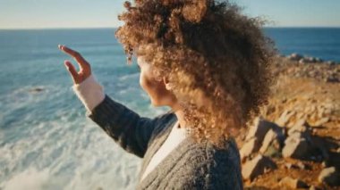 Rüzgarlı deniz kıyısında güneş ışığını ısıtan kıvırcık kadın. Rahatlamış mutlu bayan kameraya bakıyor ve sahil portresinin tadını çıkarıyor. Romantik model