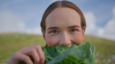 Woman holding lettuce leaf in front of face closeup. Smiling lady examining kale looking camera on green farmland field. Happy eco conscious gardener with organic vegetables. Healthy diet nutrition clipart