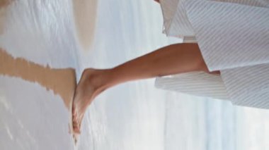 Kadın ayakları deniz sahiline dikey olarak yaklaşıyor. Okyanus kıyısında yürüyen ince bacaklar. Tanınmayan oyuncu kız elbisesiyle kumda ayak izleri bırakıyor. Rahatlamış bir kadın kıyı tatilinin tadını çıkarıyor.