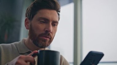 Yakın plan odaklı iş adamı geniş pencereli bir dairede elinde cep telefonu tutarak espresso içiyor. Ciddi bir adam lüks bir dairede kahve yudumluyor. Sakallı adam akıllı telefon e-postalarını okuyor.